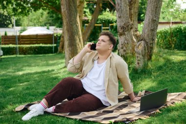 Eşcinsel bir insan parkta bir battaniyeye yaslanır, telefonla konuşur ve çalışır..