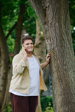 A happy person engages in a phone conversation while leaning against a tree in a park. clipart