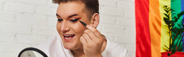 A queer individual with a big smile enjoys applying colorful makeup in a lively setting.