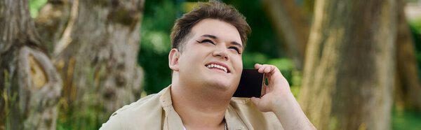 A joyful queer individual converses on the phone, surrounded by lush greenery and warm sunlight.