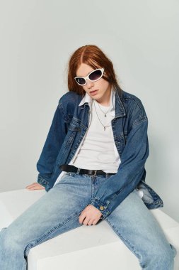 Uzun kızıl saçlı genç bir çocuk moda stüdyosunda otururken kot kıyafetini sergiliyor..