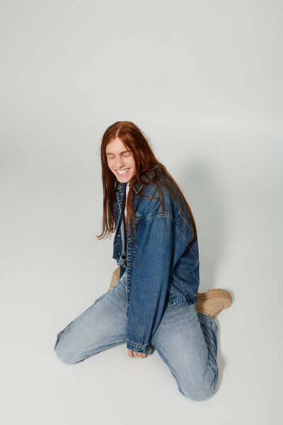 A teenage boy sits playfully on the floor, showcasing his long red hair and casual denim attire in a bright studio.