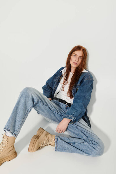 Мальчик-подросток с длинными рыжими волосами уверенно позирует в стильной джинсовой одежде на нейтральном фоне.