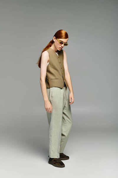 Стильный подросток с рыжими волосами уверенно стоит в умной повседневной одежде на сером фоне.