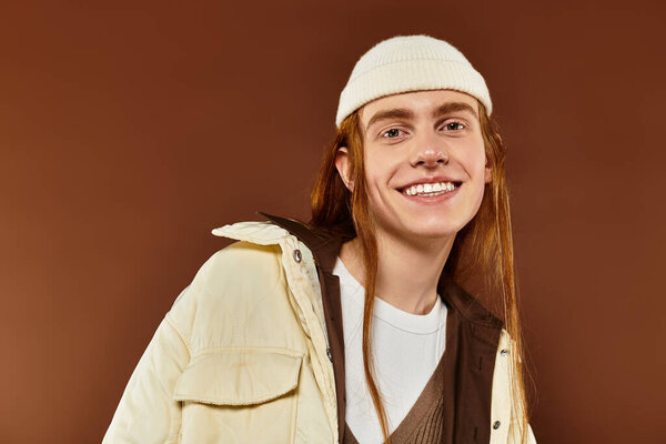 A stylish teenage boy with striking red hair smiles while posing in a cozy studio environment.