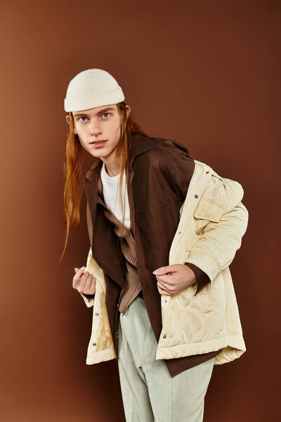 Мальчик-подросток с яркими рыжими волосами уверенно позирует в модном наряде на теплом фоне.