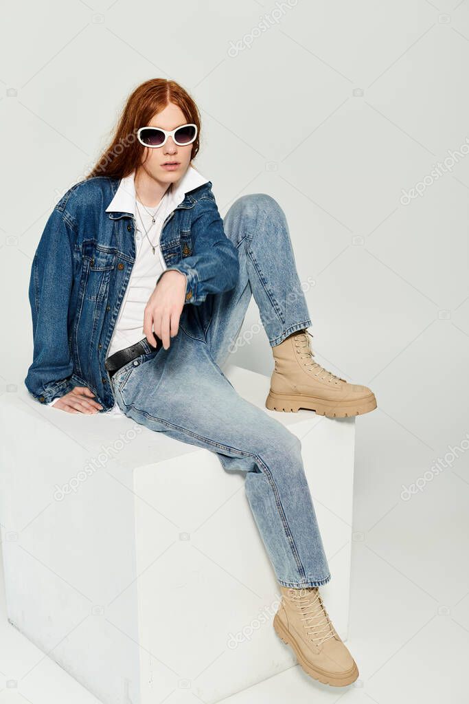 A young man with striking red hair showcases his denim style while seated on a pedestal in a studio setting.