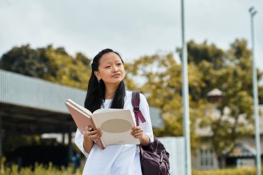 A young Asian student in a trendy outfit stands outdoors, reading her notebook with a thoughtful expression. clipart