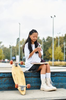 Moda düşkünü genç bir Asyalı öğrenci bir çıkıntının üzerinde rahat bir şekilde telefonunu açık hava ortamının ortasında meşgul ediyor..
