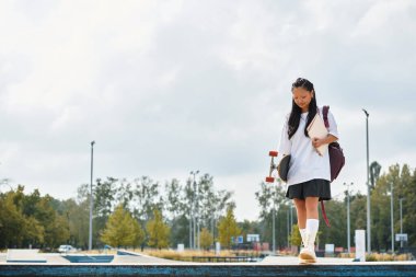 Genç ve güzel bir Asyalı öğrenci kaykayıyla parkta çeşitliliği ve gençlik kültürünü kucaklayarak yürüyor..