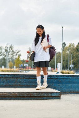 Moda kıyafetli neşeli bir öğrenci kız canlı bir parkta kaykayıyla güvenle yürüyor..