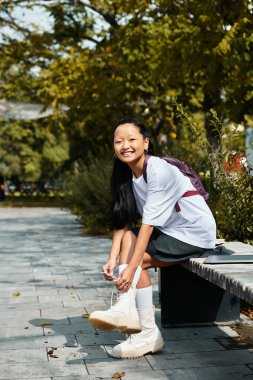 Neşeli genç bir kız bir bankta oturuyor, beyaz botlarını bağlıyor açık havada canlı bir gülümsemeyle..