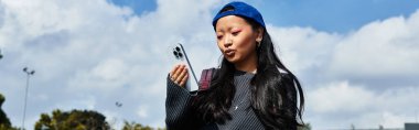 A stylish Asian student relaxes outside, absorbed in her smartphone, enjoying the vibrant day and atmosphere. clipart