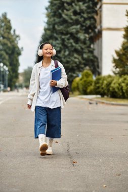 With a bright smile, a stylish young student walks through her university campus holding books under clear skies. clipart