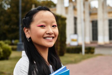 Şık bir öğrenci, üniversitesinin yanında kitap taşırken kendinden emin bir şekilde gülümsüyor, çeşitliliği ve gençliği somutlaştırıyor..