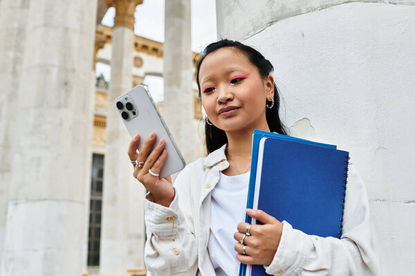 Молодая азиатская студентка в модном наряде занимается телефоном возле исторической университетской архитектуры.
