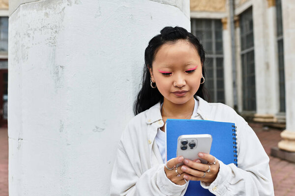 Молодая женщина проверяет телефон, держа блокнот за пределами университета, олицетворяя юношескую энергию..