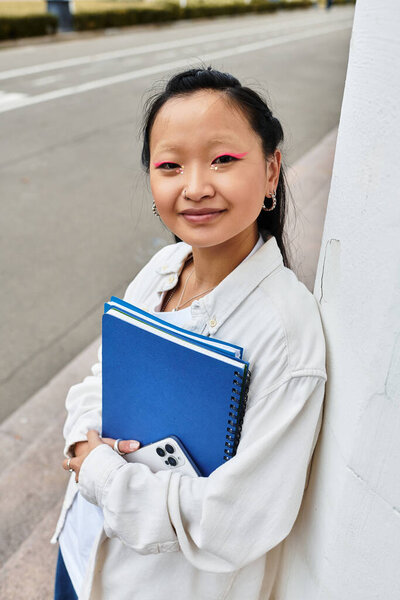 Весёлый азиатский студент прислоняется к стене, держа в руках блокноты и телефон, отражая бурную жизнь кампуса.