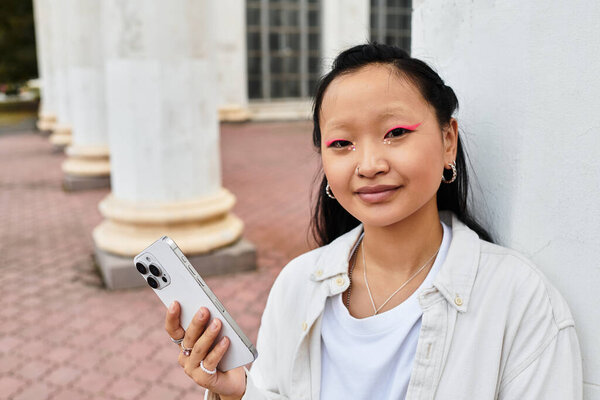 Стильная студентка уверенно стоит с телефоном, обнимая молодежную культуру за пределами университетского городка.