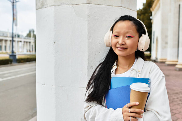 Веселая студентка уверенно стоит рядом со своим университетом, держа книги и кофе в наушниках.