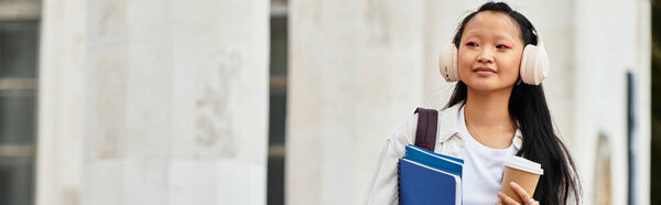 Молодой азиатский студент прогуливается по университетскому кампусу, излучая уверенность и стиль, наслаждаясь кофе.