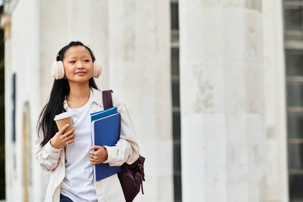 A young Asian student in a fashionable outfit walks with coffee and textbooks, embracing campus life vividly.