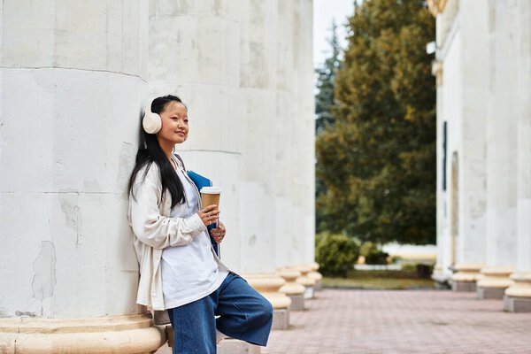 Молодой азиатский студент в наушниках улыбается, прислонившись к колонне, держа в руках кофе и книги в кампусе.