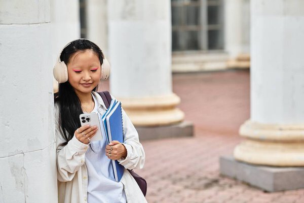 A stylish young Asian student enjoys her university campus, happily checking her phone with books in hand.