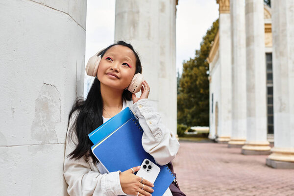 A fashionable young woman relaxes with headphones, holding notebooks and a phone in a university setting.