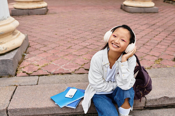 Радостная молодая азиатка в стильном наряде сидит перед университетом, наслаждаясь яркой атмосферой.