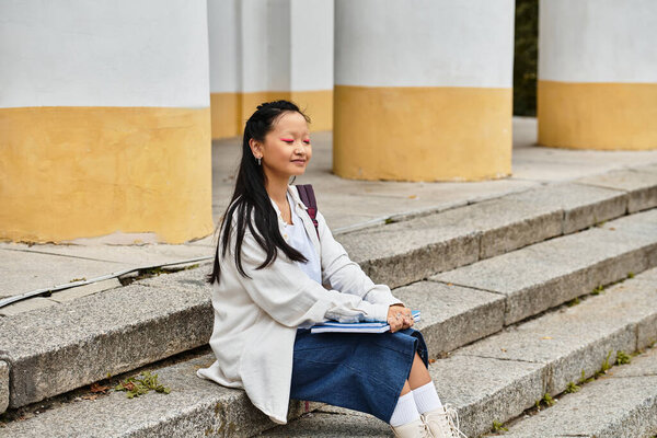 Молодая азиатская студентка отдыхает на ступеньках кампуса, принимая разнообразие и молодежную культуру в своем стильном наряде.