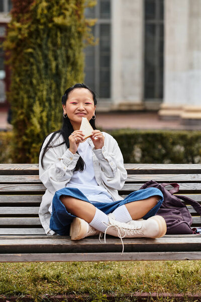 Студентка сидит на скамейке в университете, наслаждаясь сэндвичем, обнимая свой стильный наряд.