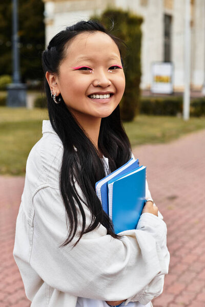 Радостная молодая азиатская студентка позирует с блокнотами, демонстрируя свой модный стиль возле университетских зданий.