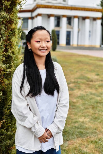 Молодая азиатская студентка в модном костюме стоит рядом со своим университетом, улыбаясь и обнимая студенческую жизнь.