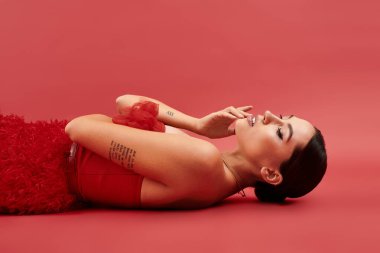 Genç bir kadın zarif bir şekilde kırmızı bir zeminde uzanır, büyüleyici tarzını sergiler ve özgüvenini sergiler..