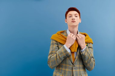 Kareli ceketli ve sarı kazaklı kendine güvenen genç bir adam stüdyo ortamında yaratıcı bir poz veriyor..