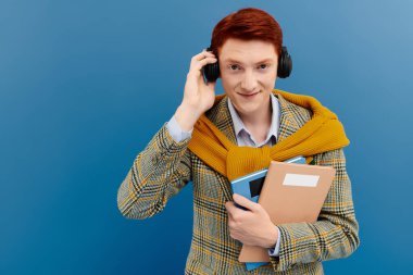 A confident young man with red hair wears an elegant layered outfit and smiles while holding books and headphones. clipart