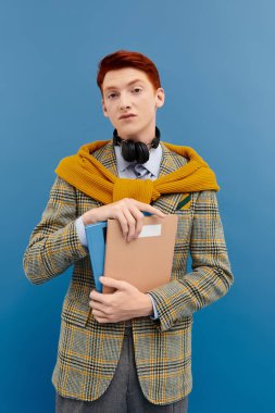 Şık bir öğrenci, mavi bir arka planda katmanlı kış kıyafeti giymiş, kendine güvenen kitapları tutar..