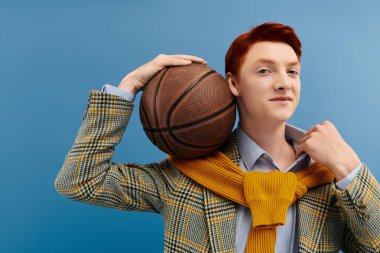 Kızıl saçlı genç bir adam şık katmanlı bir kıyafet ve kendine güvenen bir basketbol topunu stüdyoda tutuyor..