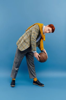 Kızıl saçlı şık bir genç, elinde basketbol topu tutarken moda anlayışını sergiliyor..