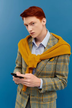 Kızıl saçlı genç bir adam mavi arka planda şık katmanlı bir kış kıyafetiyle poz veriyor..