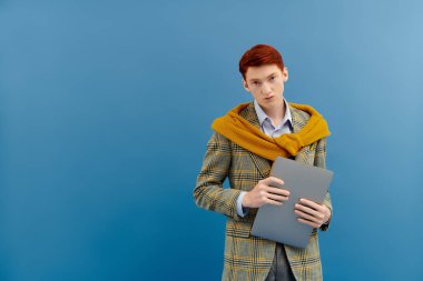 A young man with striking red hair wears a fashionable blazer and sweater, holding a gray folder in a studio. clipart