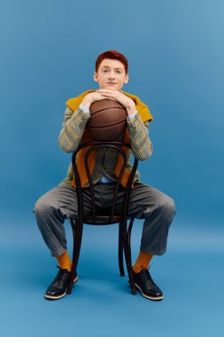 Katmanlı kış kıyafeti giyen kendine güvenen genç bir adam stüdyoda otururken bir basketbol topunu dengeliyor..