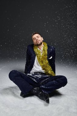 A cheerful young man dressed in a suit relaxes with a festive garland while snow falls around him. clipart