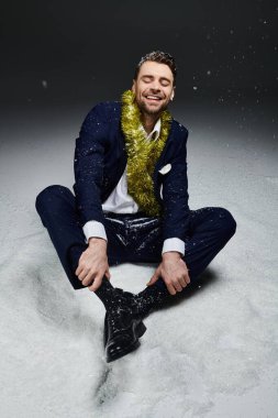 The young man laughs joyfully while sitting in the snow, dressed in a suit and festive decor. clipart