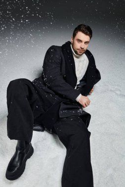 A young man reclines on the snow covered ground, dressed warmly while snowflakes fall around him. clipart