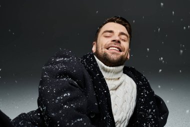 Etrafında kar taneleri yağarken yakışıklı bir genç adam geniş bir gülümseme sergiliyor ve neşeli bir atmosfer yaratıyor..