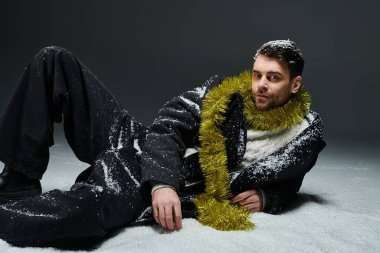 A fashionable young man relaxes on snow, adorned with festive decorations in a winter setting.