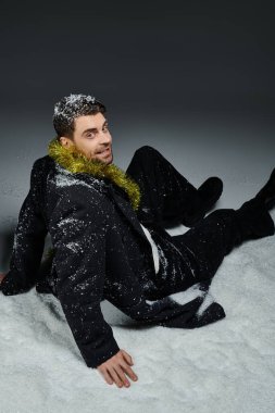 Genç adam kış eğlencesinin ortasında neşeli bir çelenkle karda oturuyor..