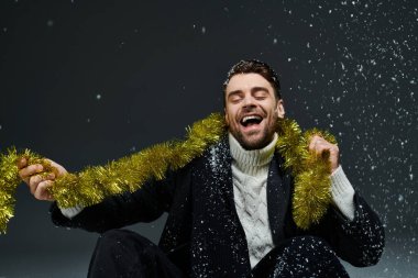 A handsome young man smiles widely as he plays with shiny tinsel amidst falling snowflakes. clipart